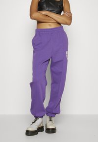 Fialové tepláky s elastickým pasem a manžetami u kotníků. Mají boční švy a grafický potisk na stehně.