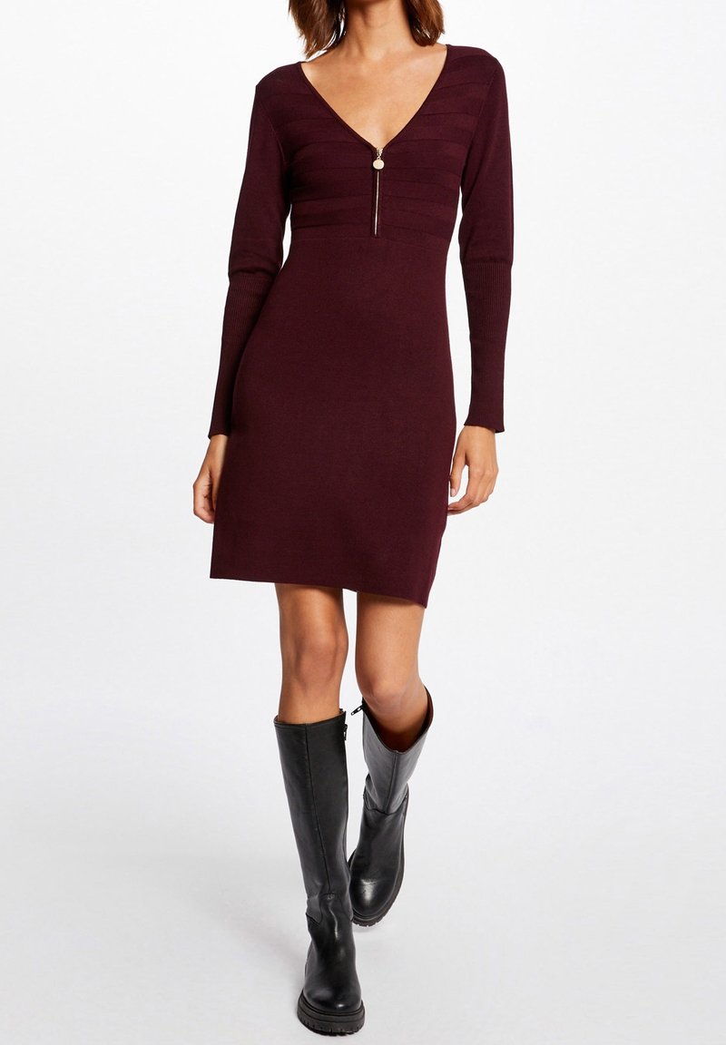 Femme portant une robe en maille bordeaux foncé à manches longues et fermeture éclair devant, avec des bottes noires jusqu’aux genoux, debout sur un fond blanc.