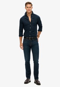 Donkerblauwe overhemden met knopen in een slim fit, gecombineerd met donkere spijkerbroek. Een zwarte leren riem en bruine loafers maken de outfit compleet.