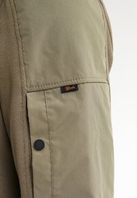 Chaqueta verde oliva con una tela texturizada, paneles lisos, una pequeña etiqueta con el logo en la manga izquierda y un detalle de botón a presión negro.