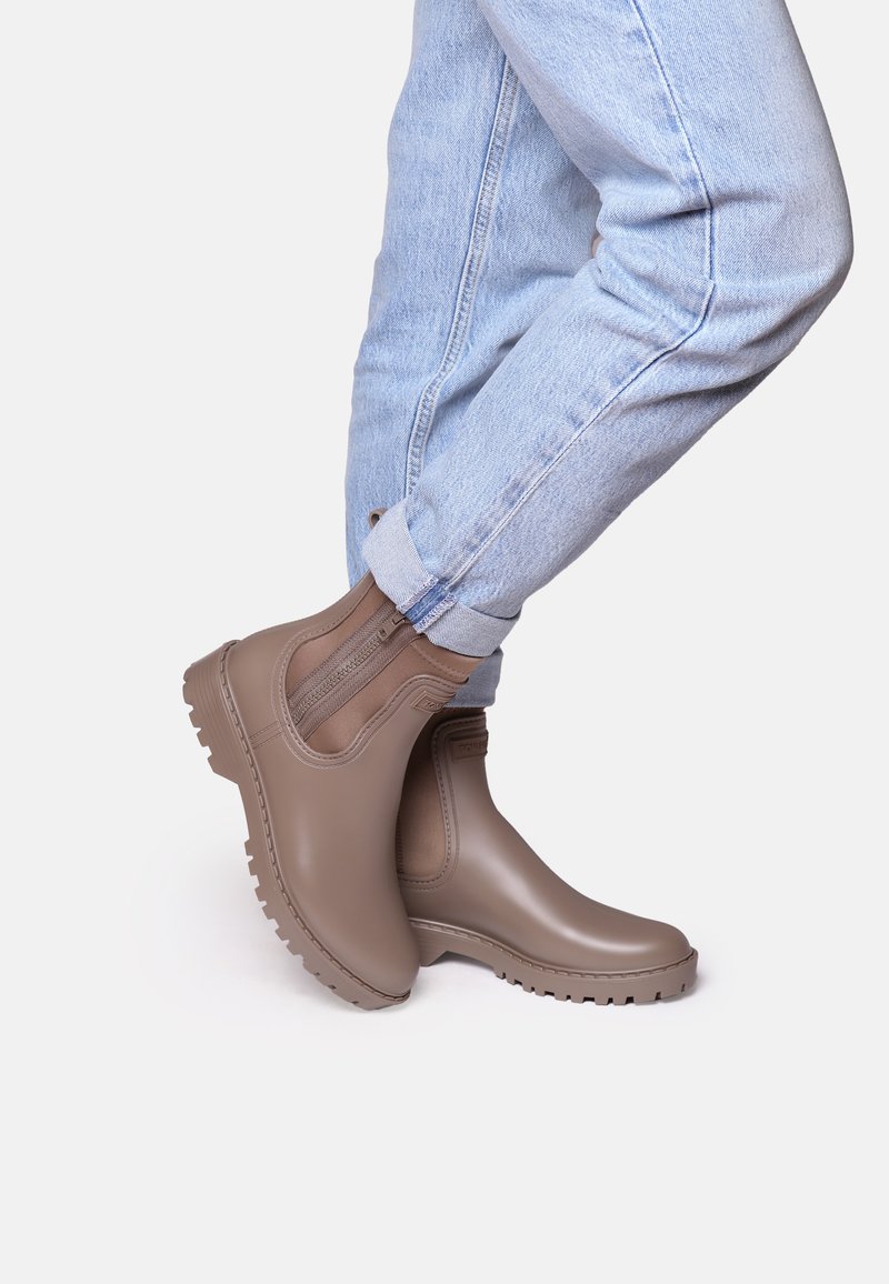 Botas de tobillo impermeables marrones con paneles laterales elásticos, suelas texturizadas y un acabado suave, combinadas con jeans azul claro arremangados.
