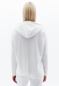 Sudadera blanca hecha de tejido texturizado, con una capucha grande, hombros caídos y un dobladillo recto. Diseño minimalista sin acentos.