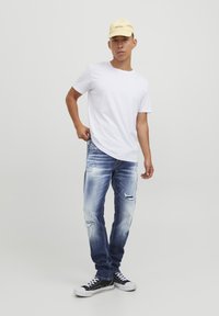 T-shirt blanc à col rond, jeans en denim bleu usé avec des zones décolorées et des déchirures, baskets noires avec semelles et lacets blancs, casquette jaune.