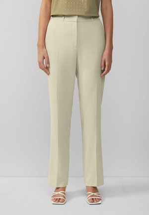 Pantaloni beige su misura con design a gamba dritta, caratterizzati da una piega anteriore precisa, passanti per cintura e una texture liscia. Abbinati a sandali bianchi con cinturini.