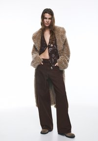 Brun fluffig lång rock över en paljettprydd mörk topp, kombinerad med vida mörkbruna byxor och ljusbruna stövlar. Texturerade material är framträdande.