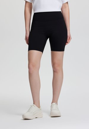 Frau trägt schwarze, hoch taillierte Biker-Shorts und weiße klobige Sneaker, steht vor einem schlichten hellen Hintergrund.