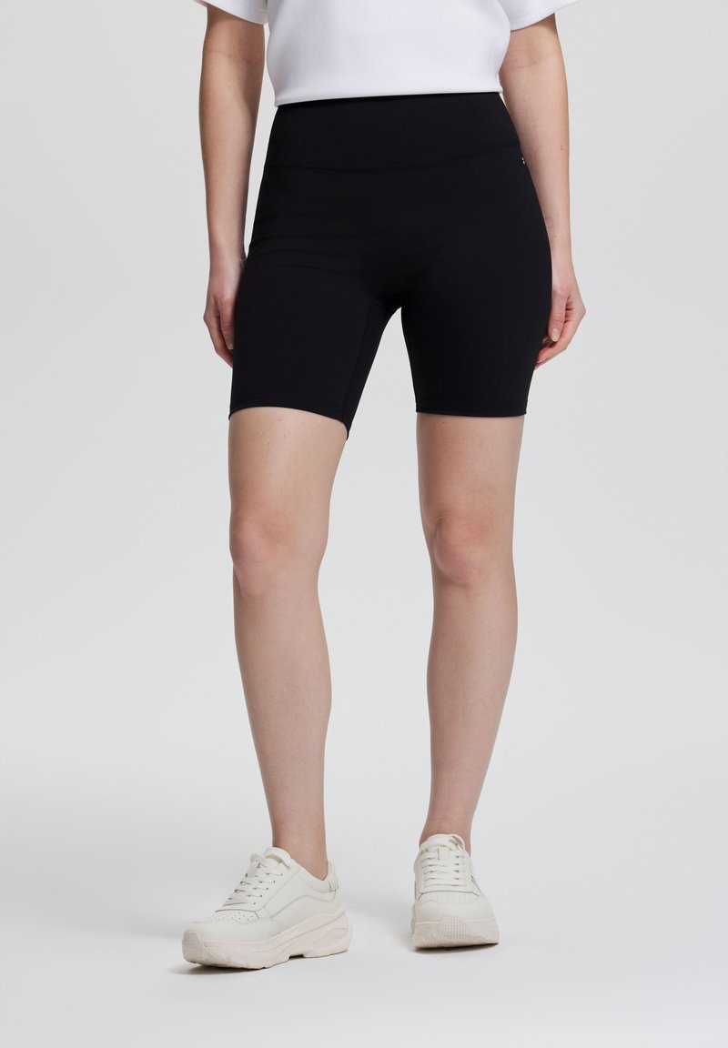 Femme portant un short de cycliste noir taille haute et des baskets blanches épaisses, debout devant un fond clair uni.