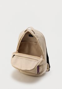 Μπεζ σακίδιο πλάτης με ανοιχτό κύριο διαμέρισμα και μπροστινή τσέπη με φερμουάρ, που διαθέτει μωβ τσόφλια φερμουάρ και μαύρους ιμάντες ώμου από δίχτυ.