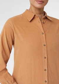 Camicia abbottonata in corduroy marrone chiaro, con colletto, maniche lunghe e una sottile texture a coste, completa di bottoni marroni lungo la parte frontale.