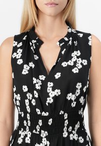 Rochie neagră fără mâneci cu un model floral alb, având un decolteu în formă de V și o talie gathered pentru o ajustare lejeră. Textură de țesătură netedă.