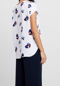 Blusa bianca morbida con motivi astratti blu, rossi e navy, maniche corte, scollatura rotonda e orlo curvato. Tessuto morbido.