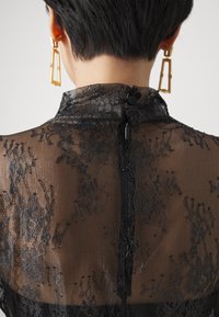 Blusa de encaje negro con cuello alto, textura transparente, patrones florales y un cierre en la parte posterior con un ojal y dos botones. Pendientes geométricos dorados.