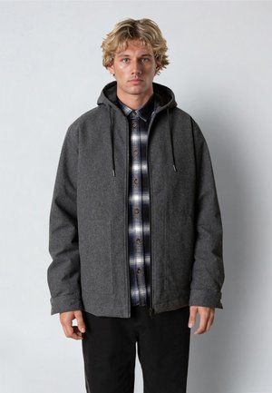 Ung mand med blondt hår iført en grå hættetrøje over en sort og hvid ternet skjorte, stående mod en ensfarvet baggrund.