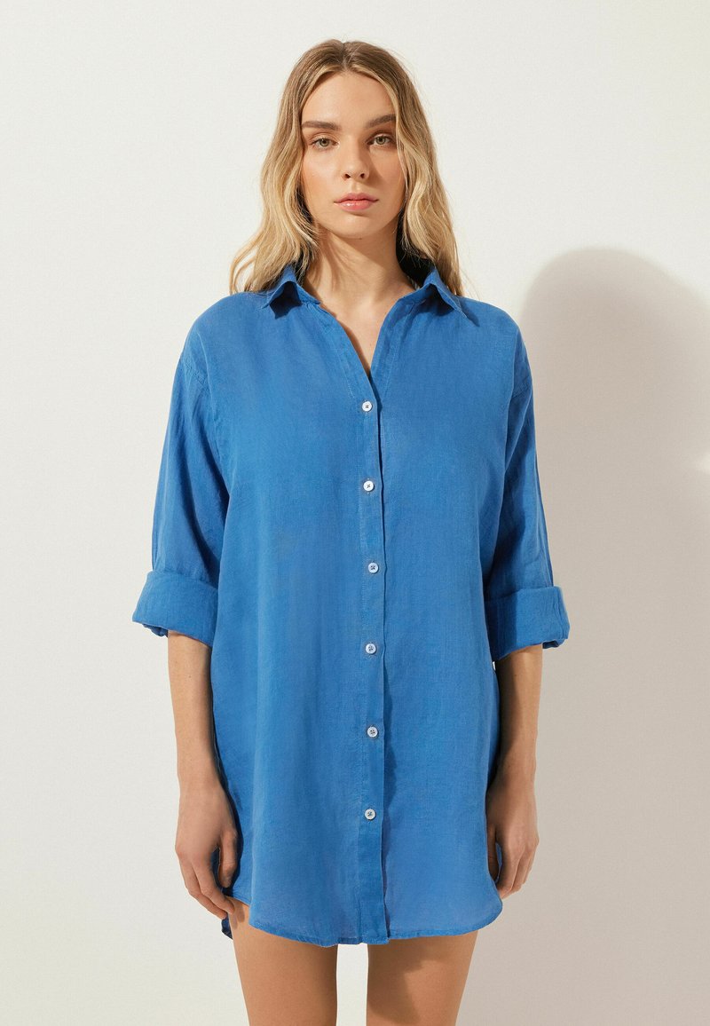 Ljusblå skjorta med knappar och avslappnad passform, uppvikta ärmar och en strukturerad väv. Har en spetsig krage och synliga knappar.