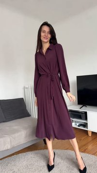 Długa sukienka zapinana na guziki w głębokim fiolecie, z wiązanym paskiem w talii, długimi rękawami i zwiewnym dołem. Noszona z czarnymi szpilkami.