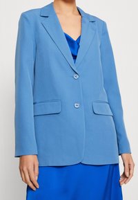 Modrý blazer se dvěma předními kapsami, zapínáním na jeden knoflík a klopovými límci, vyrobený z hladké látky, vrstvený přes modrou top. Kn.