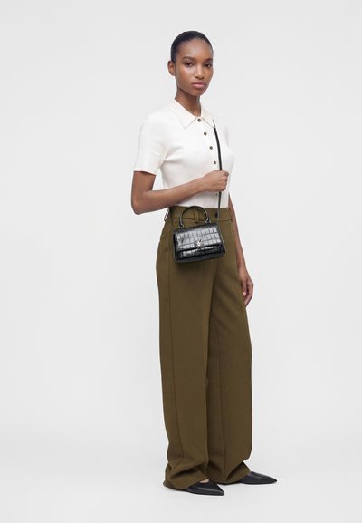 Femme portant un haut blanc à manches courtes et un pantalon large olive, tenant un petit sac à main noir texturé avec une bandoulière.
