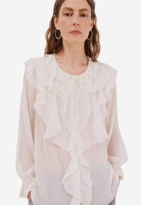 Bílá halenka s volánkovým předkem, šňůrkou na zavazování v oblasti výstřihu a dlouhými rukávy. Vyrobená z lehké látky s texturovaným povrchem.