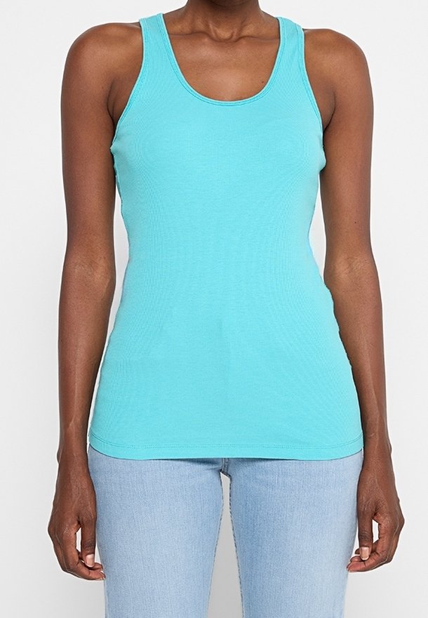 Femme portant un débardeur turquoise sans manches et un jean bleu clair, debout devant un fond blanc uni.