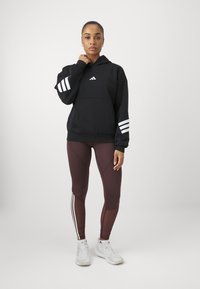Sweatshirt preto com design de três riscas brancas nas mangas, bolso canguru, combinado com leggings bordôs com listras laterais. Ténis brancos.