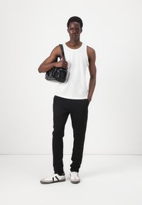 Geantă din piele lacuită neagră cu un design prins, purtată peste un maieu alb cu dungi și pantaloni negri slim fit, asortată cu adidași gri și albi.