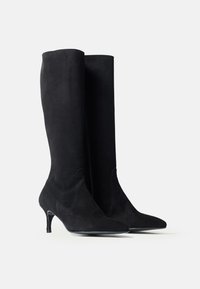 Botine negre din suede, până la genunchi, cu vârf ascuțit și toc subțire de înălțime medie, expuse una lângă alta pe un fundal alb simplu.