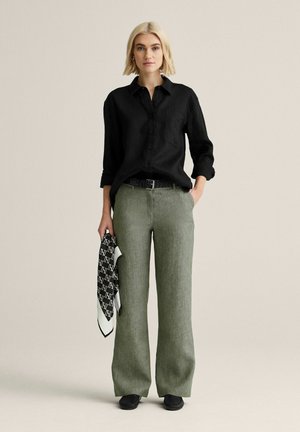 Lyshåret kvinde iført sort skjorte med knapper, olivengrønne vidde bukser, sort bælte, sorte sko, holder sort-hvidt, mønstret tørklæde i højre hånd.