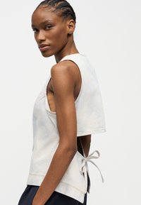 Femme aux cheveux tressés portant un haut sans manches rayé clair avec des liens sur les côtés, légèrement tournée de profil sur un fond uni.