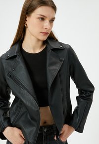 Zwarte faux leren motorjack met een ritssluiting aan de voorkant, notch-kraag, drukknoopjes en ritsmouwen. G gedragen over een zwart getailleerd top.