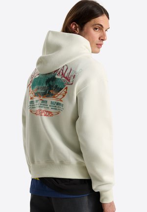 Jonge man met lang haar die zich omdraait, draagt een off-white hoodie met een kleurrijke afbeelding en tekst op de achterkant.