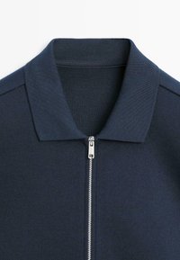 Marineblå jakke med lynlås og ribbet krave. Glat tekstur og rene linjer, designet til et tætsiddende udseende.