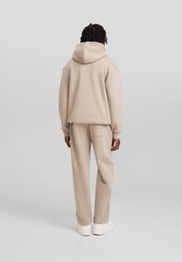 Felpa beige con cappuccio e vestibilità ampia, abbinata a pantaloni coordinati. Tessuto liscio, con una tasca sul retro dei pantaloni.