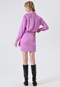 Rosa Satinlangarmhemd kombiniert mit einem Minirock. Das Model trägt schwarze kniehohe Stiefel. Sanfte Textur, taillierte Passform, minimale Details.