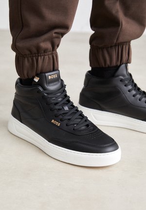 Baskets montantes noires avec semelles blanches et marquage doré "BOSS", portées avec un pantalon de jogging marron sur un sol clair.