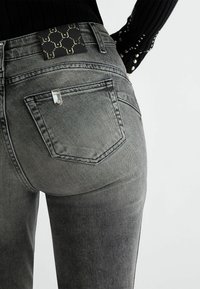 Jeans de mezclilla gris con un acabado texturizado, que cuentan con bolsillos traseros y una etiqueta de marca negra con un patrón repetido cerca de la cintura.