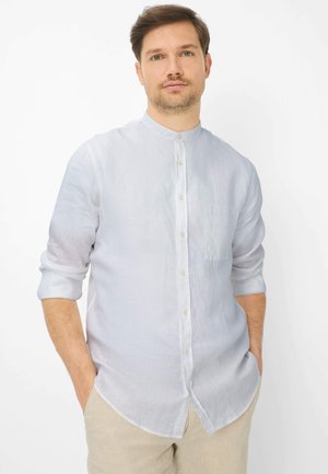 Mann mit hellbraunem Haar, der ein weißes Mandarin-Kragenhemd mit hochgekrempelten Ärmeln und beige Hosen trägt, die Hände in den Taschen, neutraler Hintergrund.