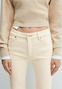 Jean clair à taille haute, avec poches avant et fermeture à bouton en métal. Associé à un pull court beige. Texture lisse.