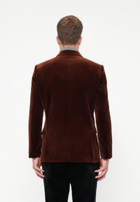 Vyras atsisukęs atgal, vilkintis tamsiai rudą aksominį švarką, po kuriuo pilkas polo megztinis ir juodos kelnės, priešais vientisą baltą foną.