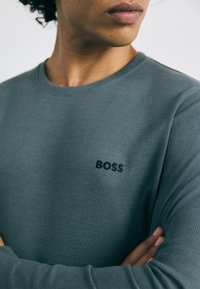 Tmavý teal dlhý rukáv textúrovaná mikina s okrúhlym výstrihom a rebrovaním. Obsahuje tónový vyšívaný logo "BOSS" na hrudi.