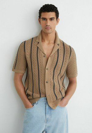 Junger Mann mit lockigem Haar, der ein beiges kurzärmeliges Strickshirt mit schwarzen vertikalen Streifen und hellblaue Jeans trägt, steht mit den Händen in den Taschen.