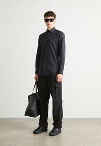 Camisa negra de botones, pantalones cargo negros, zapatillas negras y una bolsa de tote negra. Material suave, ajuste estructurado y diseño minimalista.