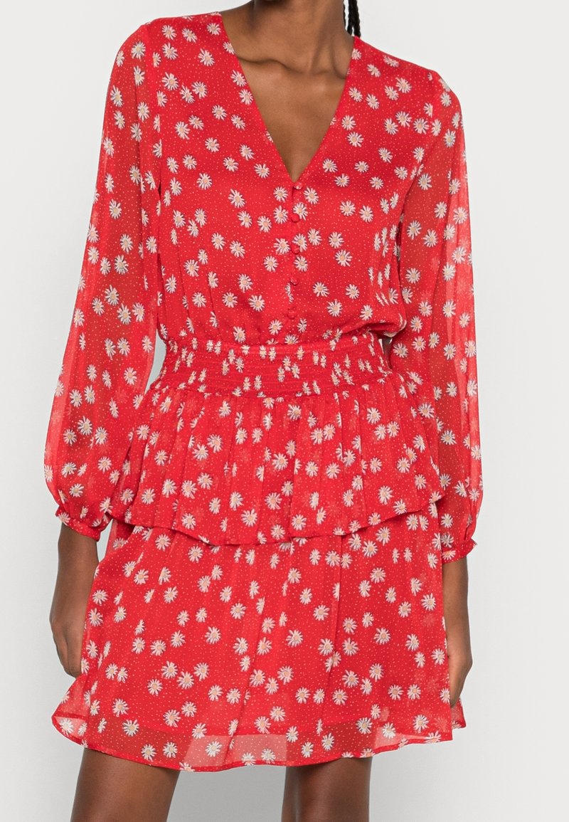 Robe rouge à manches longues avec imprimé marguerite blanche, encolure en V, taille cintrée et jupe à volants portée par une personne debout.