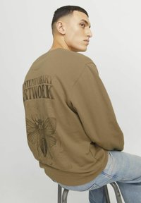 Sudadera marrón con una ilustración de una polilla y el texto "OBRA DE ARTE CONTEMPORÁNEA" en la parte trasera, combinada con jeans de color azul claro.