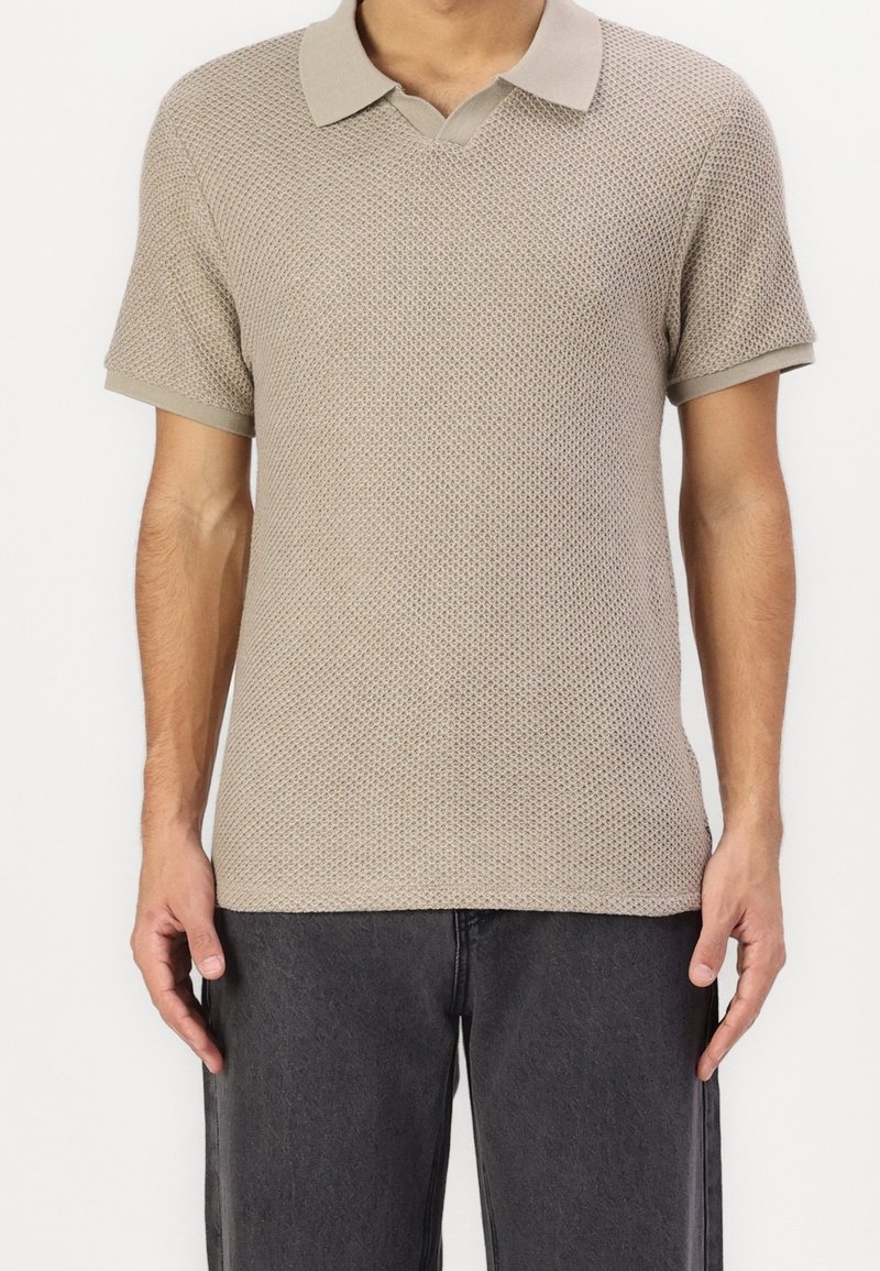 Polo en tricot beige à manches courtes et col assorti, présentant un motif texturé et un design en v.