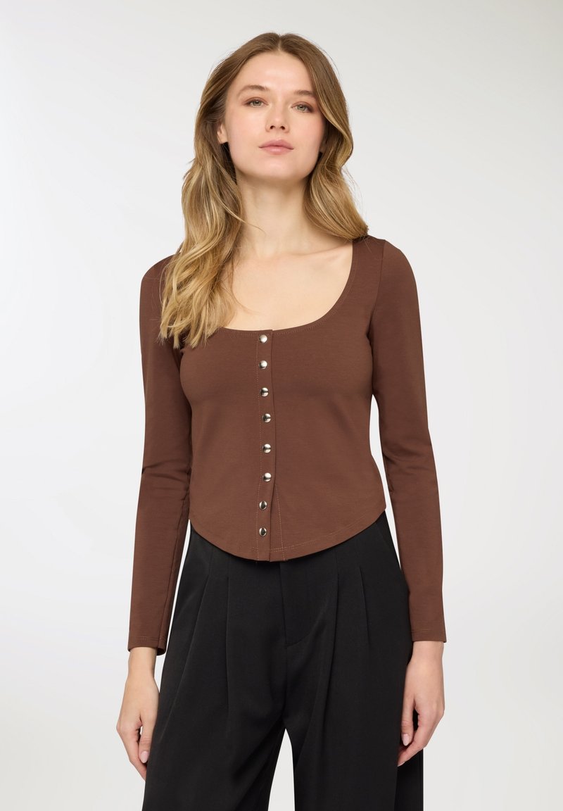 Maglietta marrone a maniche lunghe con scollo ampio e chiusura frontale con bottoni. Presenta un design aderente e un orlo corto, abbinata a pantaloni neri.