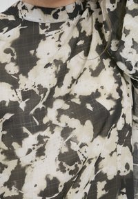 Tessuto a motivi neri e beige con una texture fluida, caratterizzato da un design astratto a macchie e un collo alto con dettagli a griglia visibili.