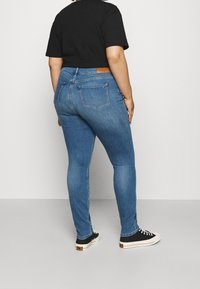 Chude niebieskie dżinsy z całkowicie średnim stanem, z brązową skórzaną metką z tyłu, pięcioma kieszeniami i gładką teksturą denimu.