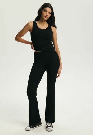 Kobieta o długich, ciemnych włosach, ubrana w czarny top na ramiączkach, czarne rozkloszowane spodnie i czarno-białe trampki, stojąca z jedną ręką na biodrze.