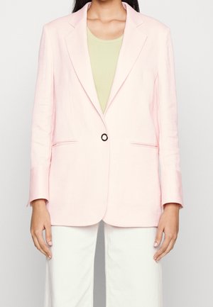 Kvinde iført lyserød blazer med enkelt knap over en lysegrøn top og hvide bukser, stående mod en ensfarvet baggrund.