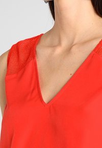 Rød ærmeløs top med V-hals design og teksturerede mesh-nuancer på skuldrene. Glat stof med en afslappet pasform.